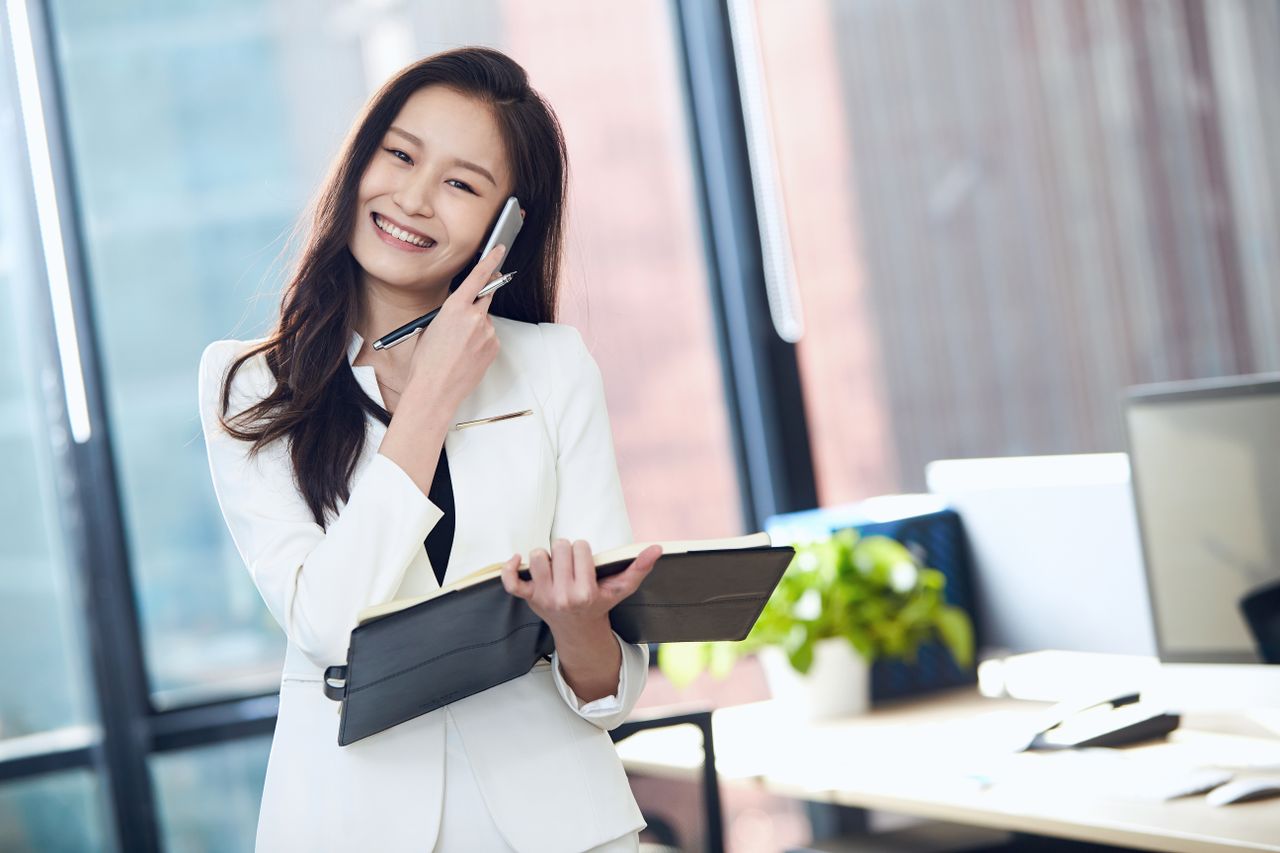 Confident Asian businesswoman in white suit, smiling and talking on mobile phone in a bright office.
